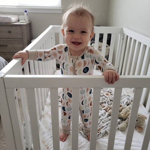 Smiling baby standing in a crib wearing the Luna Neutral Zippy footie