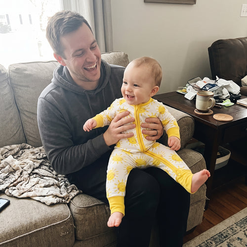 Dad laughing with his baby on the couch in Sweet Sleep Sunshine Zippy pajamas