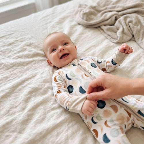 Smiling baby on a bed wearing the Luna Neutral Zippy moon print zip footie