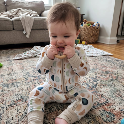 Baby sitting on a rug chewing a teether in the Luna Neutral Zippy footie