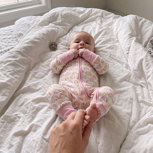 Baby lying on a bed wearing the Sweet Sleep Pastel Rainbows Zippy footie pajamas